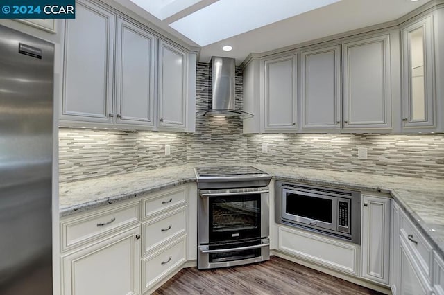 kitchen featuring hardwood / wood-style floors, wall chimney range hood, a skylight, appliances with stainless steel finishes, and tasteful backsplash