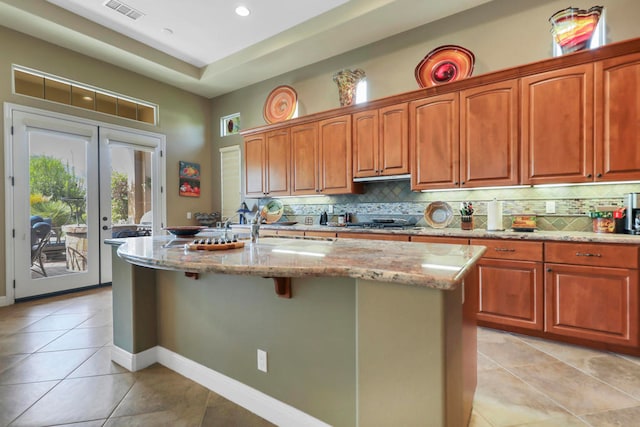 kitchen with decorative backsplash, light tile patterned flooring, light stone counters, stainless steel gas stovetop, and a center island with sink