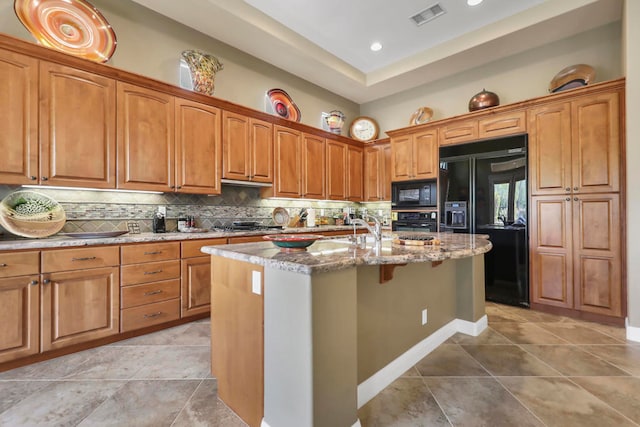 kitchen featuring decorative backsplash, light stone countertops, light tile patterned floors, black appliances, and a center island with sink