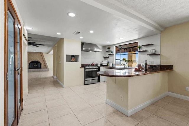 kitchen featuring wall chimney range hood, stainless steel range oven, kitchen peninsula, a fireplace, and light tile patterned floors