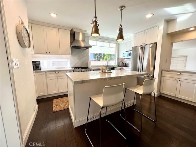 kitchen featuring decorative light fixtures, wall chimney range hood, a kitchen island, a breakfast bar, and appliances with stainless steel finishes