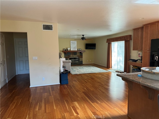 kitchen with black fridge with ice dispenser, dark wood-type flooring, a stone fireplace, and ceiling fan