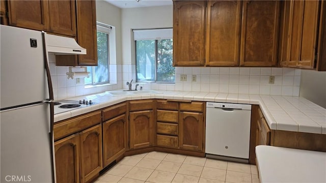 kitchen with white appliances, light tile patterned floors, backsplash, under cabinet range hood, and a sink