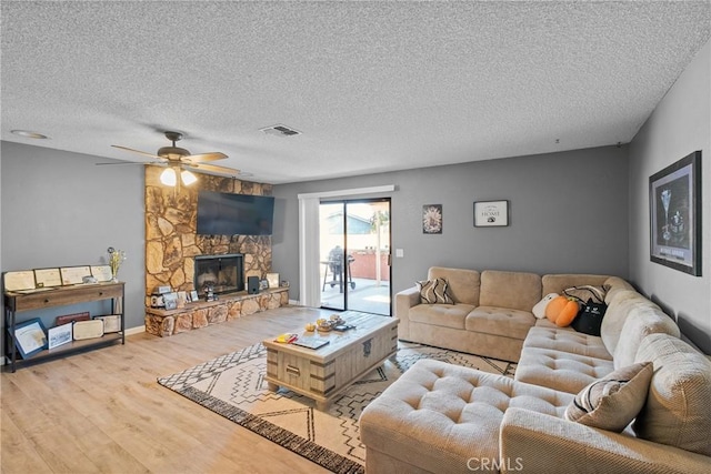 living room featuring a textured ceiling, light wood-type flooring, a stone fireplace, and ceiling fan