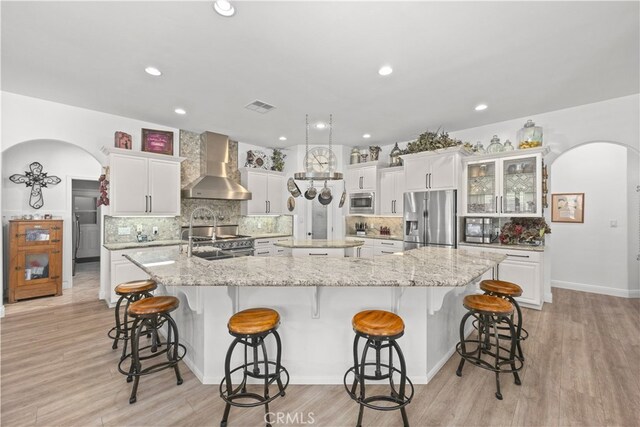 kitchen with a large island, white cabinets, wall chimney range hood, light hardwood / wood-style flooring, and appliances with stainless steel finishes