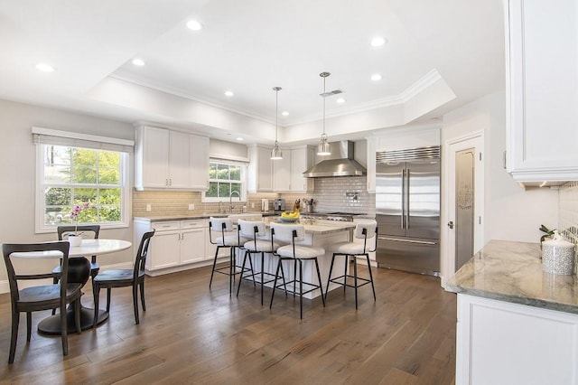 kitchen with a tray ceiling, wall chimney exhaust hood, built in fridge, and white cabinetry