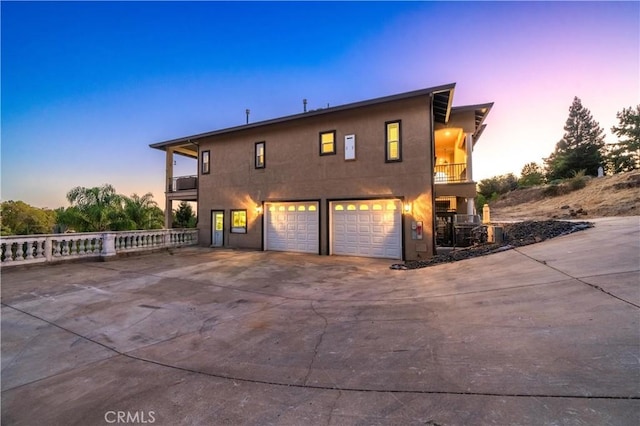 back of property at dusk featuring stucco siding, an attached garage, a balcony, and driveway