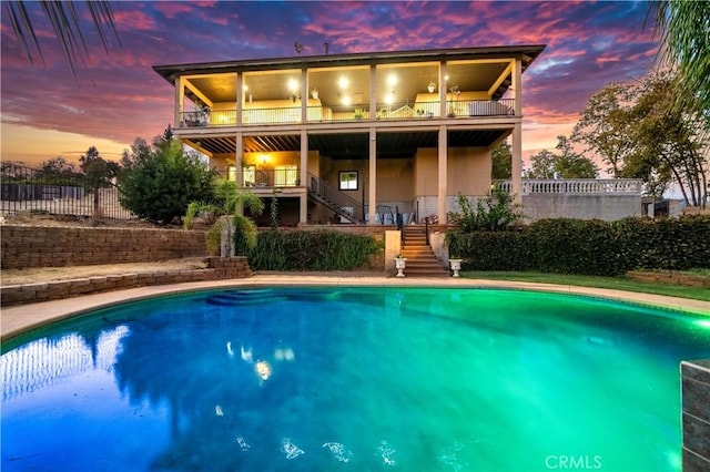 back of property at dusk with a fenced backyard, a balcony, stairs, and a fenced in pool
