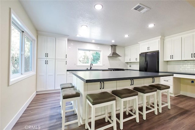 kitchen with white cabinetry, dark wood-type flooring, wall chimney range hood, black fridge, and a breakfast bar area