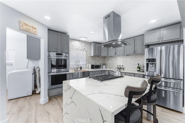 kitchen with light wood-type flooring, sink, island exhaust hood, stainless steel appliances, and light stone countertops