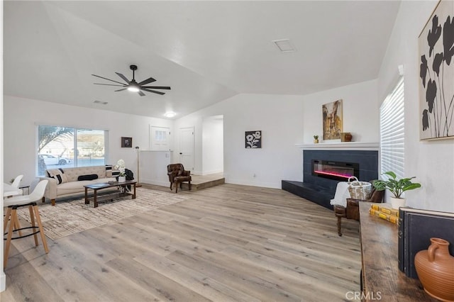 living area featuring visible vents, a glass covered fireplace, ceiling fan, wood finished floors, and vaulted ceiling