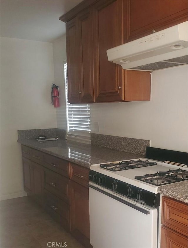 kitchen featuring dark stone counters, white range with gas stovetop, brown cabinets, and under cabinet range hood