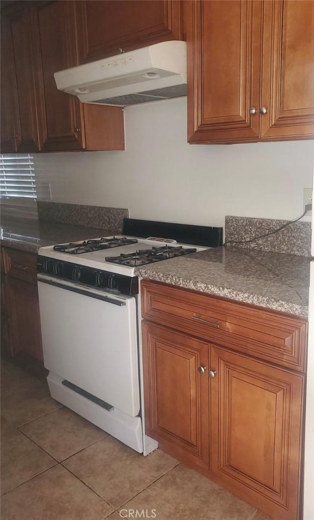 kitchen featuring brown cabinetry, light tile patterned flooring, white gas stove, and under cabinet range hood