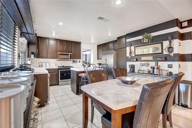 kitchen featuring backsplash, dark brown cabinetry, stainless steel appliances, light tile patterned floors, and a kitchen island