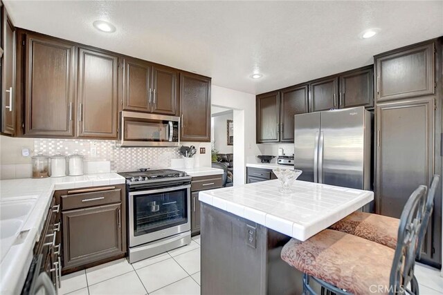 kitchen with tile countertops, a kitchen breakfast bar, light tile patterned floors, appliances with stainless steel finishes, and dark brown cabinetry