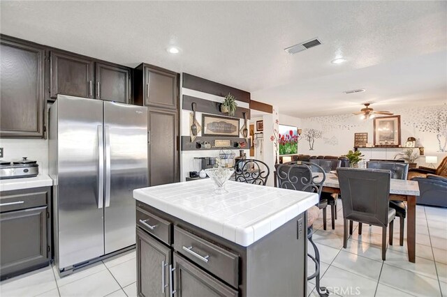 kitchen with tile countertops, ceiling fan, light tile patterned floors, a kitchen island, and stainless steel refrigerator