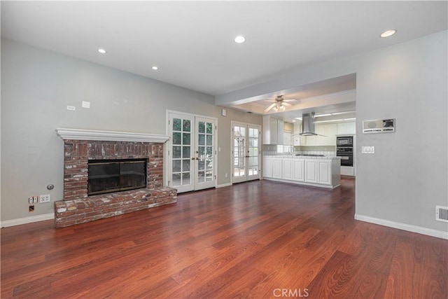 unfurnished living room featuring a brick fireplace, ceiling fan, dark wood-type flooring, and french doors