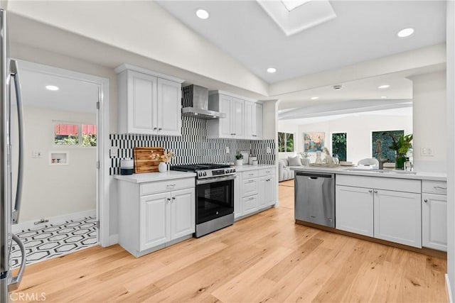 kitchen with wall chimney exhaust hood, lofted ceiling with skylight, white cabinetry, light wood-type flooring, and appliances with stainless steel finishes