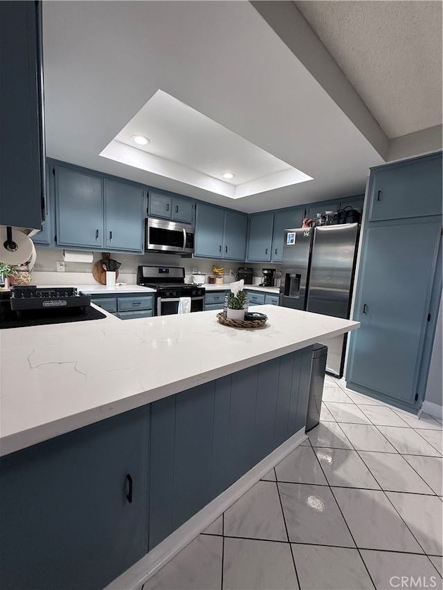 kitchen with light tile patterned floors, kitchen peninsula, blue cabinetry, appliances with stainless steel finishes, and a tray ceiling