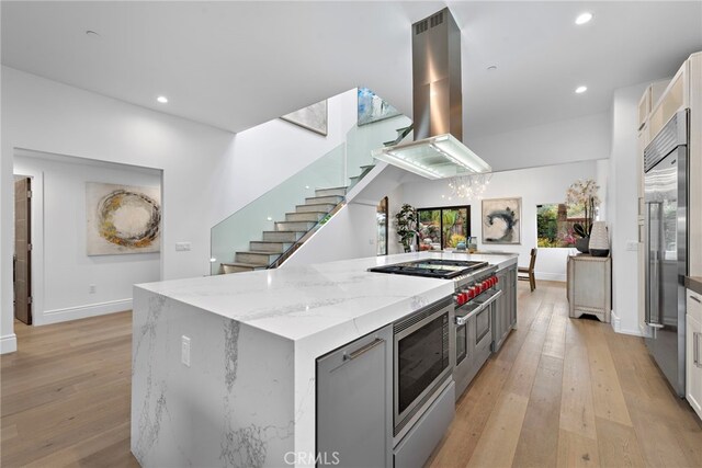 kitchen with light stone countertops, a kitchen island, island range hood, light hardwood / wood-style floors, and built in appliances