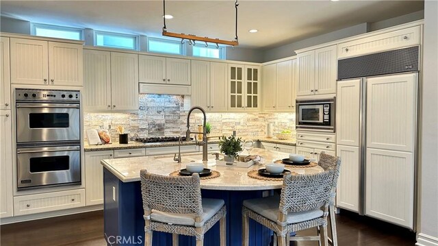 kitchen featuring decorative backsplash, light stone counters, built in appliances, decorative light fixtures, and an island with sink