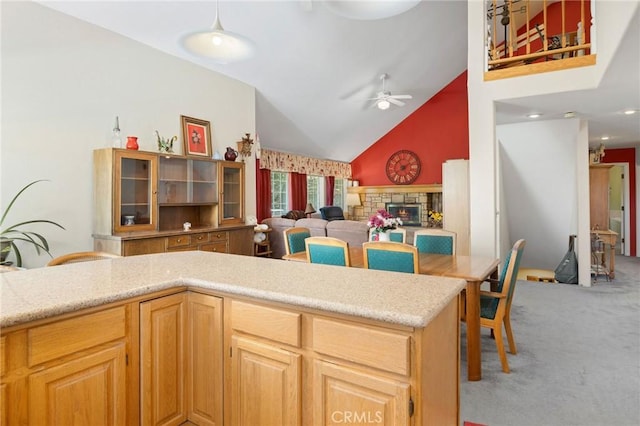 kitchen with carpet flooring, light brown cabinetry, ceiling fan, high vaulted ceiling, and a stone fireplace