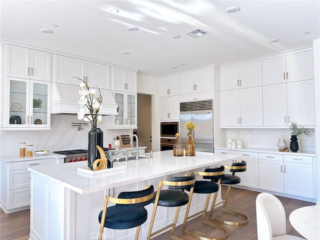 kitchen with built in appliances, white cabinets, a kitchen island with sink, and dark wood-type flooring