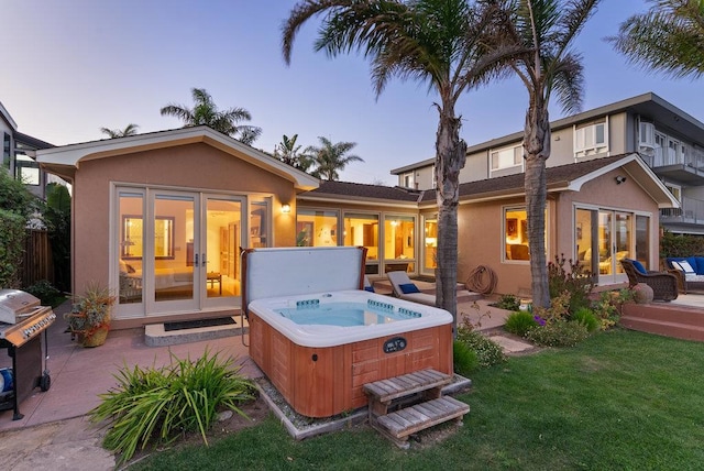 back house at dusk with a hot tub, a patio area, and a lawn