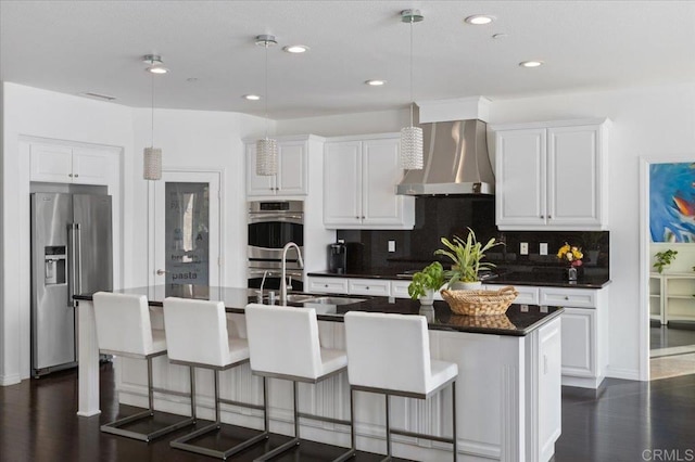 kitchen featuring a center island with sink, wall chimney range hood, appliances with stainless steel finishes, and white cabinetry