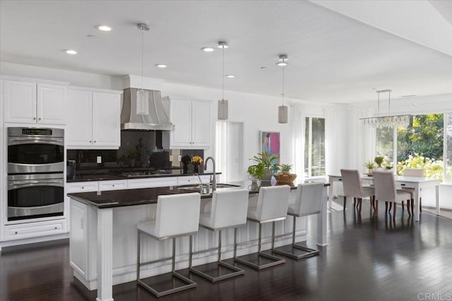 kitchen featuring hanging light fixtures, wall chimney range hood, white cabinets, and a center island with sink