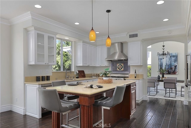 kitchen featuring pendant lighting, white cabinetry, ornamental molding, a center island, and wall chimney range hood