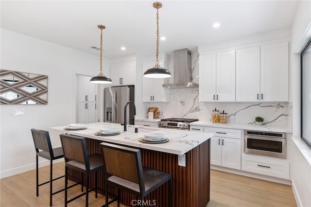 kitchen with white cabinets, light hardwood / wood-style floors, wall chimney range hood, appliances with stainless steel finishes, and a center island with sink