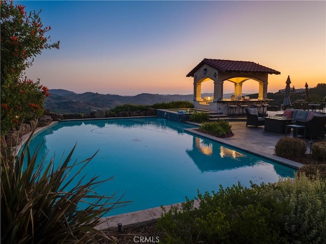 pool at dusk with an outdoor bar, a patio area, a mountain view, and an in ground hot tub