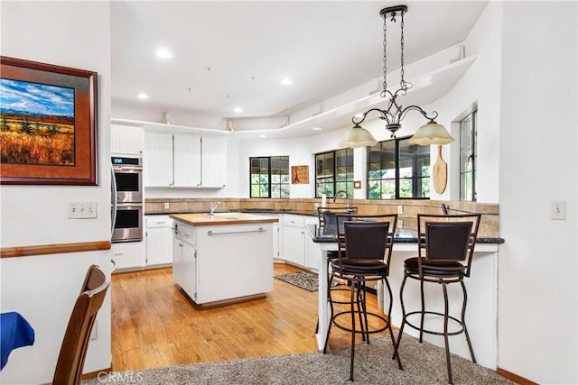 kitchen featuring white cabinetry, kitchen peninsula, stainless steel double oven, hanging light fixtures, and light hardwood / wood-style flooring