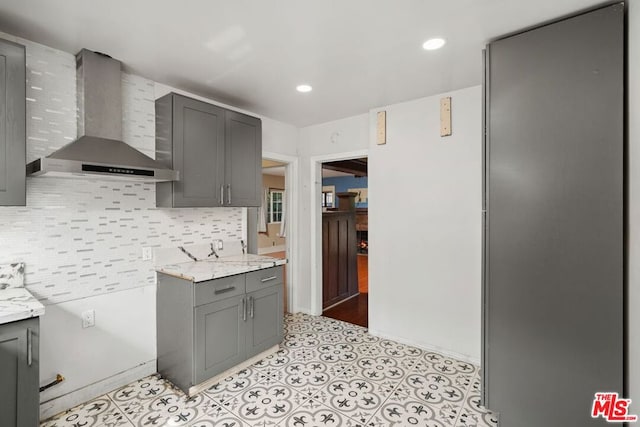 kitchen featuring gray cabinetry, decorative backsplash, wall chimney exhaust hood, and light stone countertops