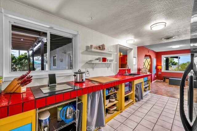 kitchen featuring tile countertops, open shelves, light tile patterned flooring, a sink, and stainless steel microwave