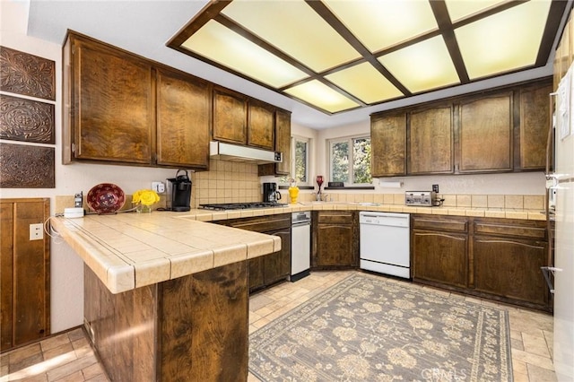 kitchen with white dishwasher, a peninsula, dark brown cabinetry, under cabinet range hood, and tile countertops