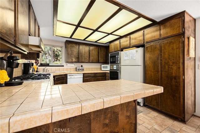 kitchen with white appliances, dark brown cabinetry, a peninsula, under cabinet range hood, and tile counters
