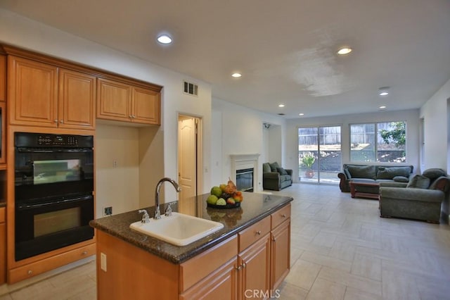 kitchen with double oven, a center island with sink, dark stone counters, and sink