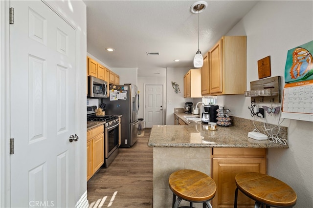 kitchen with kitchen peninsula, dark hardwood / wood-style flooring, appliances with stainless steel finishes, a kitchen bar, and light stone counters