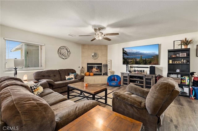 living room with a tiled fireplace, wood-type flooring, a textured ceiling, and ceiling fan