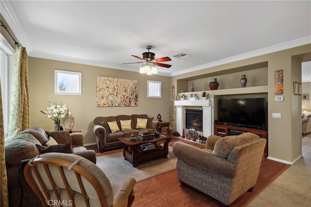 living area with wood finished floors, a glass covered fireplace, ceiling fan, ornamental molding, and visible vents