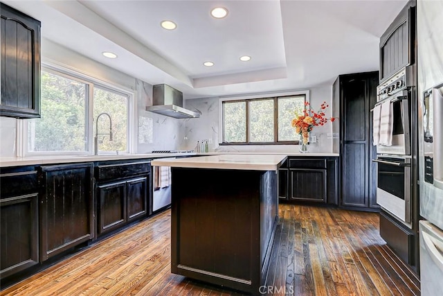 kitchen featuring dark hardwood / wood-style flooring, a tray ceiling, sink, wall chimney range hood, and a center island