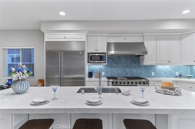 kitchen with white cabinetry, wall chimney range hood, built in appliances, a kitchen breakfast bar, and backsplash