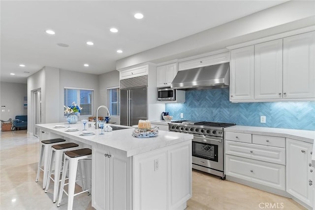 kitchen with an island with sink, built in appliances, range hood, white cabinets, and sink