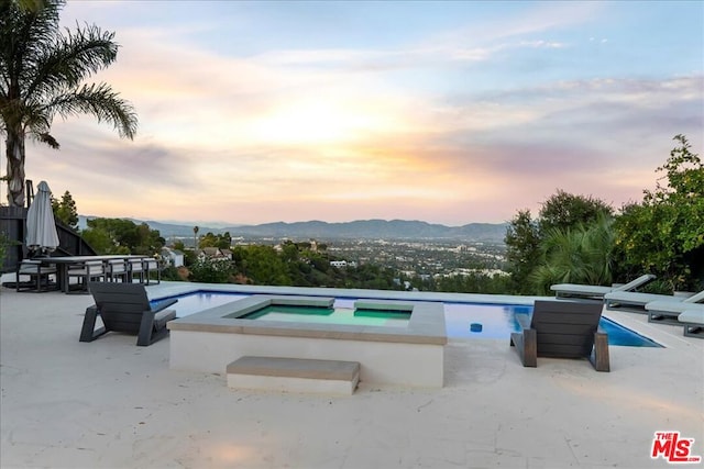 pool at dusk with an in ground hot tub, a mountain view, and a patio