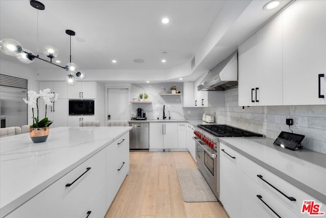 kitchen with wall chimney range hood, decorative light fixtures, white cabinetry, light wood-type flooring, and appliances with stainless steel finishes