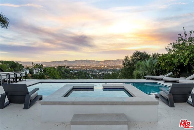 pool at dusk with an in ground hot tub, a mountain view, and a patio