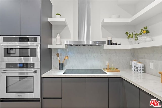 kitchen with double oven, decorative backsplash, gray cabinets, and wall chimney range hood