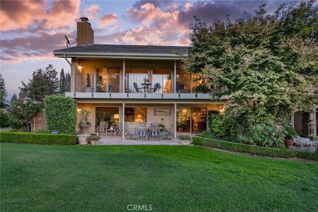 back house at dusk with a patio, a balcony, and a lawn